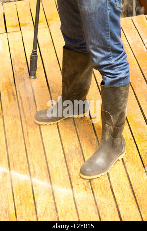 Man wearing rubber boots using high water pressure cleaner on wooden terrace surface. Stock Photo