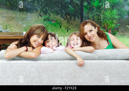Two proud hispanic mothers posing with their daughters, heads sticking up from sofa and smiling to camera Stock Photo