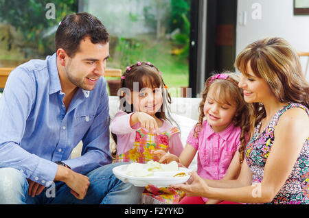 Hispanic parents with two daughters eating from a tray of potato chips sitting in sofa while smiling and enjoying each other company Stock Photo