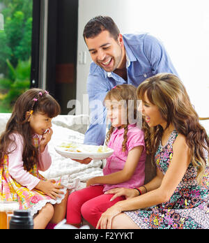 Hispanic parents with two daughters eating from a tray of potato chips sitting in sofa while smiling and enjoying each other company Stock Photo