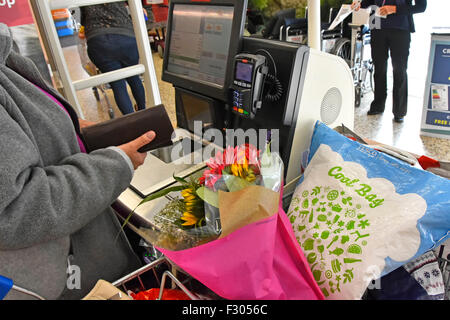 Woman shopping Tesco self service checkout scan as you shop till download data from hand held scanner beside full weekly food shop trolley England UK Stock Photo