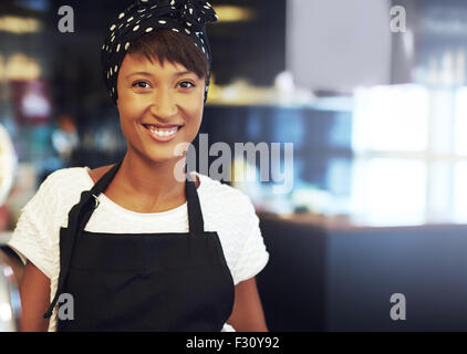 Successful young African American business owner standing in her coffee shop in an apron and bandanna smiling at the camera Stock Photo
