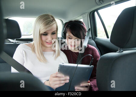 Two teen sisters in the back seat of a car holding and sharing an ipad tablet. Stock Photo