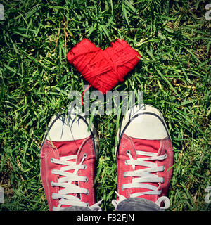 a heart-shaped coil of red rope and the feet of a man wearing red sneakers stepping on the grass, with a slight vignette added Stock Photo