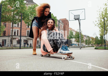 Happy young woman sitting on longboard being pushed by her friend along the road. Girls enjoying skating outdoors on city street