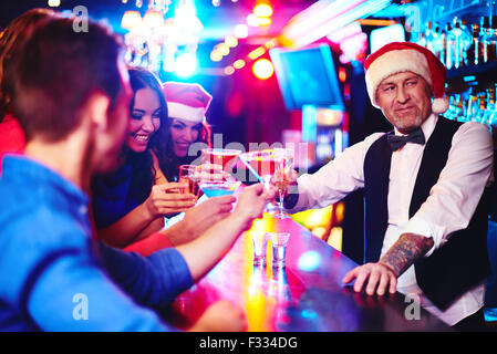 Happy company cheering up by counter with barman in Santa cap near by Stock Photo