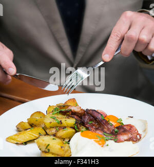 closeup of man in a suit eating fried eggs, potatoes and bacon Stock Photo