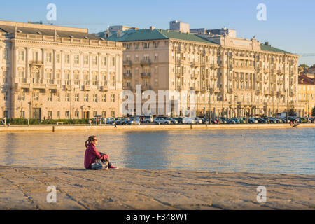 Woman thinking alone, view of a young woman sitting alone in Trieste harbour and watching the sunset on the Adriatic, Italy. Stock Photo