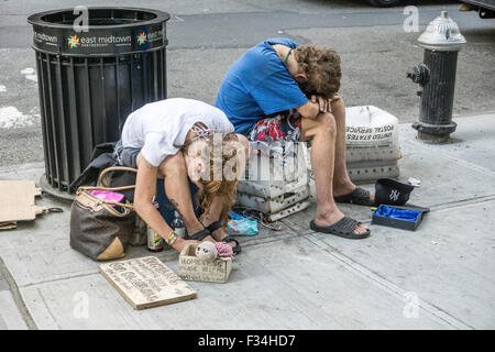 dejected homeless young people couple sit on the sidewalk in midtown Manhattan begging for spare change with heads lowered Stock Photo