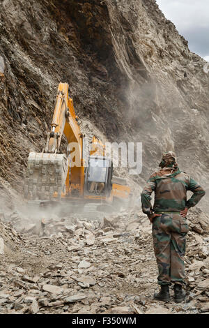 Officers from Border Roads Organization and GREF watch as excavator clears hazardous Himalayan mountain road following landslide Stock Photo