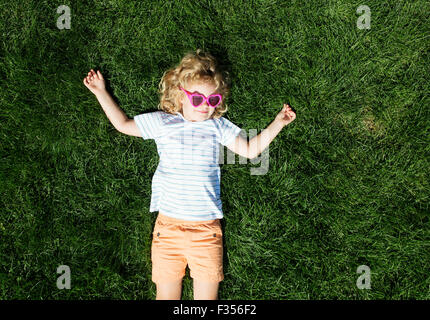 Portrait of a smiling little girl in heart shaped sunglasses lying on green grass Stock Photo