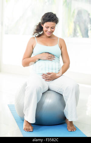 Smiling pregnant woman touching her belly while sitting on exercise ball Stock Photo