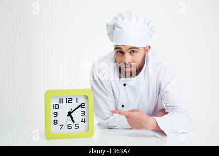 Portrait of a chef cook leaning on the table and pointing finger on clock isolated on a white background Stock Photo