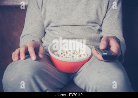 Young man is sitting on a sofa and eating popcorn while watching television Stock Photo