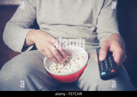 Young man is sitting on a sofa and eating popcorn while watching television Stock Photo