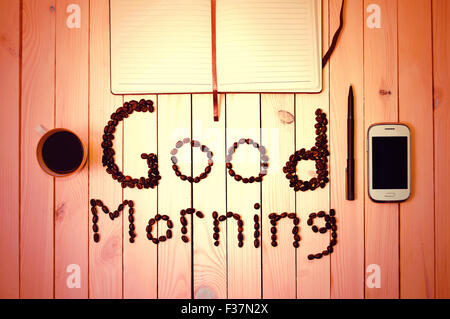Good Morning inscription made with coffee beans between white cell phone, cup of coffee, pen and notebook on wooden table. Coffe Stock Photo
