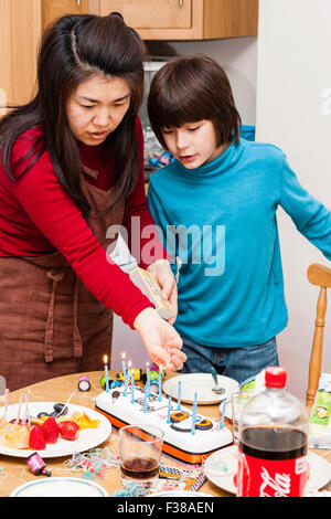 Child, boy, 9-11 year old, mixed race, Caucasian Japanese, standing while his Japanese mother prepares his birthday cake with candles on kitchen table. Stock Photo