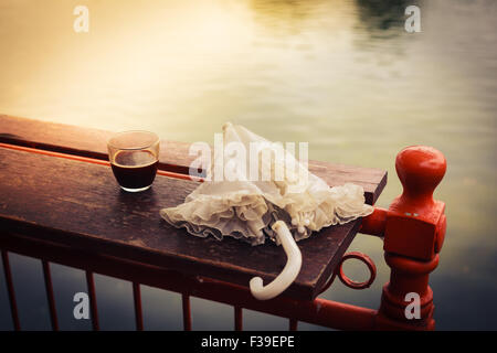 A glass of coffee and an umbrella on a wooden table by a lake in the afternoon Stock Photo