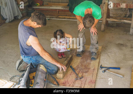 Girl helping two workmen drill a hole in a piece of  wood Stock Photo