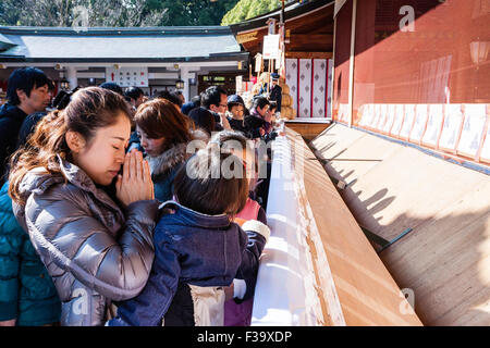 Japan, new year, winter. Japanese woman, mother, holding hands together and praying with two infant children at crowded Shinto shrine. Daytime. Stock Photo