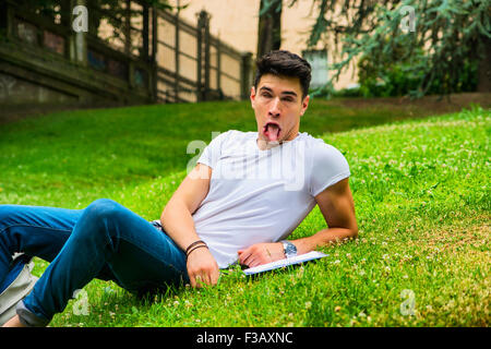Young Male Student Doing Silly Face While Studying his Lessons, Lying on Grass in City Park Stock Photo