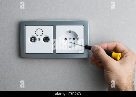 Installing a silver wall-mounted AC power socket with a screwdriver on a grey wall, renovating home. Close up view. Stock Photo