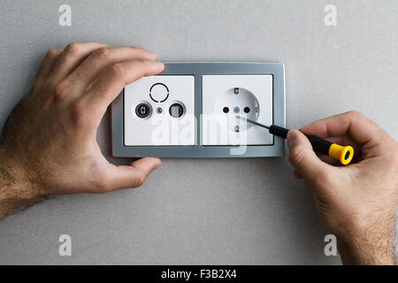 Installing a silver wall-mounted AC power socket with a screwdriver on a grey wall, renovating home. Close up view. Stock Photo