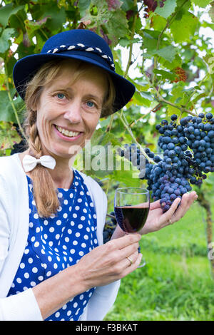 Caucasian middle aged woman holding glass of red wine near bunch of blue grapes in vineyard Stock Photo