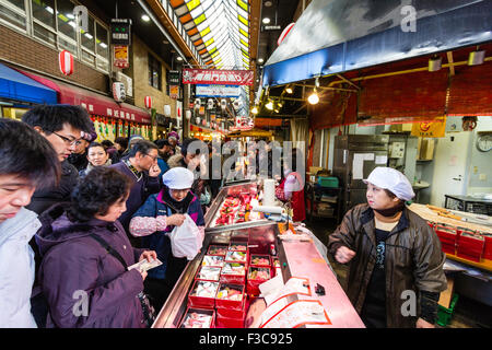 Crowds of people shopping at the busy Kuromon Ichiba market, Also known as Osaka's kitchen. People buying sushi in pre-packed boxes from counter. Stock Photo