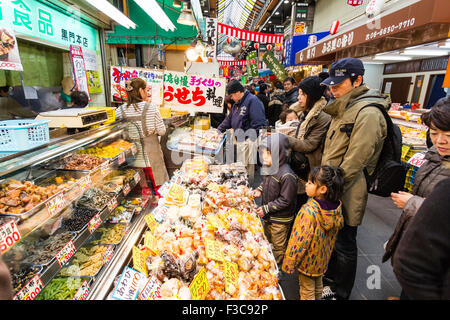 The Kuromon Ichiba Market, known as 'Osaka Kitchen' in Osaka, Japan. Customers viewing sushi and other foods on display counter at stall. Stock Photo