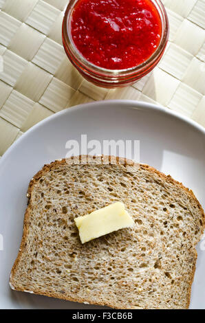 Slice of whole grain bread with pat of butter on a plate and small jar of homemade strawberry jam.  Slice of toast with butter and jar of preserves. Stock Photo