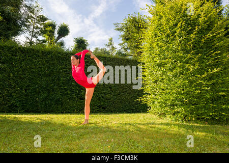 Young woman, wearing a red-orange body suit, is practising Hatha-Yoga outdoor between trees, showing the pose: natarajasana, Lor Stock Photo