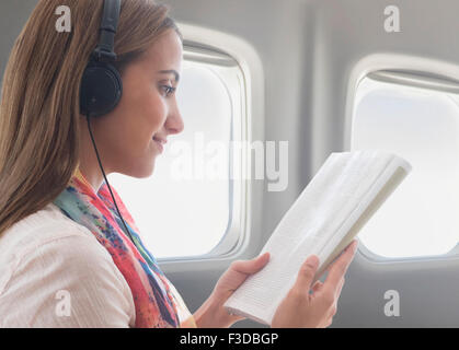 Young woman wearing headphones while reading book on plane Stock Photo