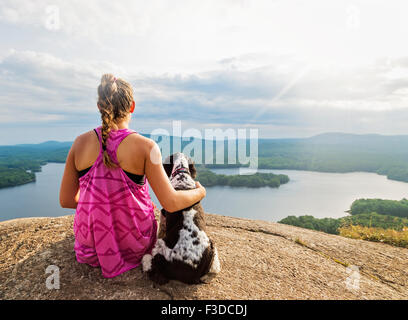 Young woman sitting with dog on cliff looking at lake view Stock Photo