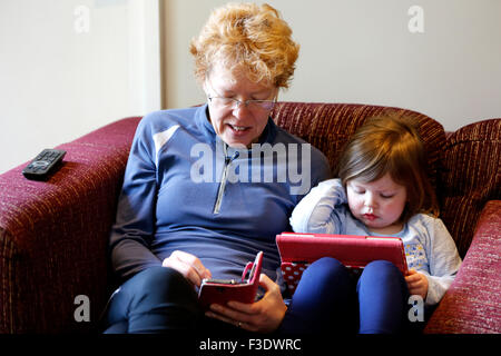 a grandmother and grandchild sit side by side on a sofa. They are using a mobile phone and an tablet device to browse the internet Stock Photo