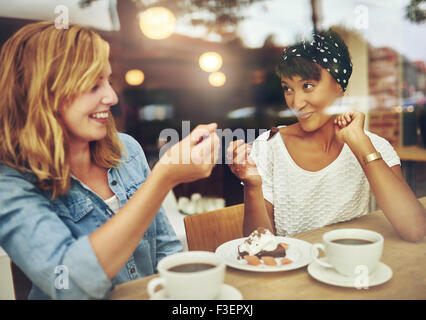 Two charismatic multi ethnic young girl friends sit at a counter in a cafe enjoying a cup of coffee while laughing and chatting Stock Photo
