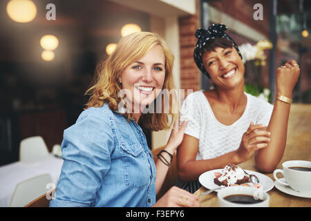 Two attractive young multiethnic female friends enjoying coffee and cake together in a cafeteria smiling happily at the camera a Stock Photo