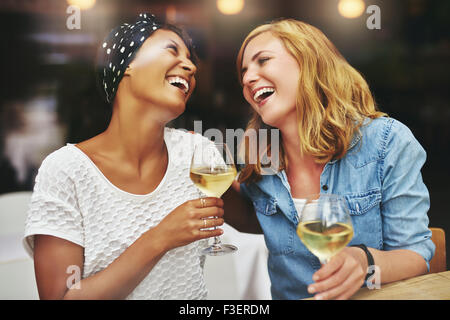 Two young attractive vivacious multiethnic female friends celebrating and laughing together over a glass of white wine Stock Photo