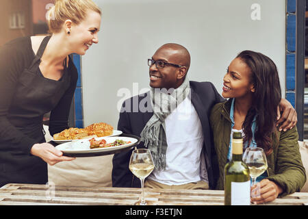 Smiling happy young Caucasian waitress serving a loving African American couple dinner as they sit arm in arm at a table in a re Stock Photo