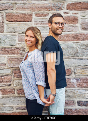 Three Quarter Shot of a Happy Young Couple Standing Back to Back While Holding their Hands and Smiling at the Camera Against Bri Stock Photo