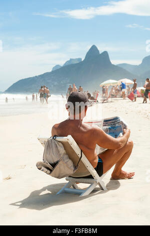 RIO DE JANEIRO, BRAZIL - JANUARY 20, 2013: Brazilian man sits sunbathing on Ipanema Beach reading a newspaper. Stock Photo