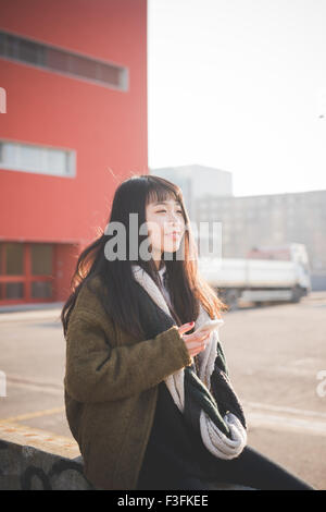 young beautiful asian hipster long brown straight hair woman sitting on a small wall, using a smartphone, overlooking left, smiling during sunset in backlight - technology, social network concept Stock Photo