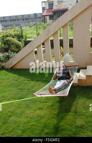 Retired person resting on hammock in bungalow garden ; Talegaon ; Pune ; Maharashtra ; India MR Stock Photo