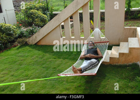 Retired person resting on hammock in bungalow garden ; Talegaon ; Pune ; Maharashtra ; India MR Stock Photo