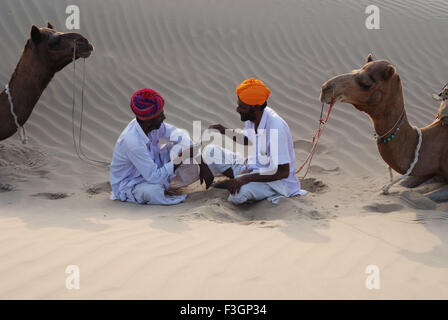 Two men with camels resting in desert ; Khuri ; Jaisalmer ; Rajasthan ; India Stock Photo