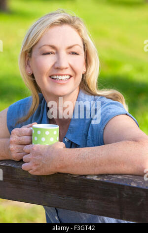 Beautiful happy middle aged woman in her thirties or forties smiling and leaning on fence in sunshine drinking tea or coffee Stock Photo