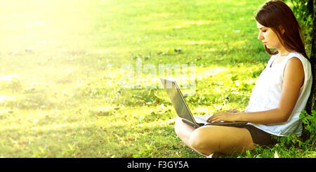 Young beautiful college student girl sitting on the green grass and working on laptop at campus at warm day. Education. Stock Photo