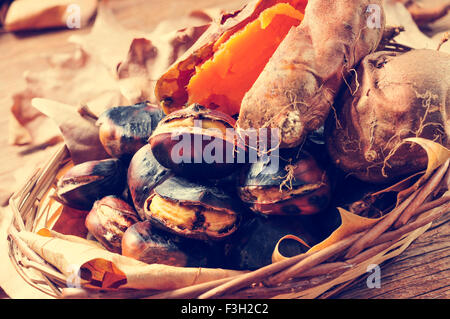 some roasted chestnuts and some roasted sweet potatoes in a wicker basket with autumn leaves, on a rustic wooden table Stock Photo