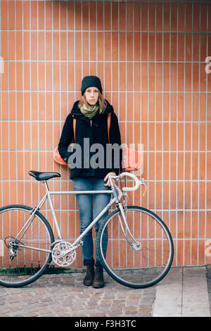 young beautiful hipster sporty blonde woman posing leaning against a wall with her bike, headphones around her chest and hand in pocket. Looking in camera. Music, sportive concept Stock Photo