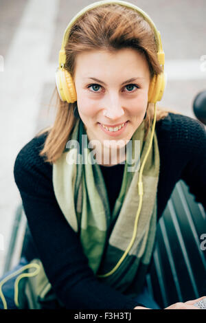 Portrait of young beautiful hipster sporty blonde woman sitting on a bench, looking in camera, smiling, listening music with headphones - music, relaxing concept Stock Photo